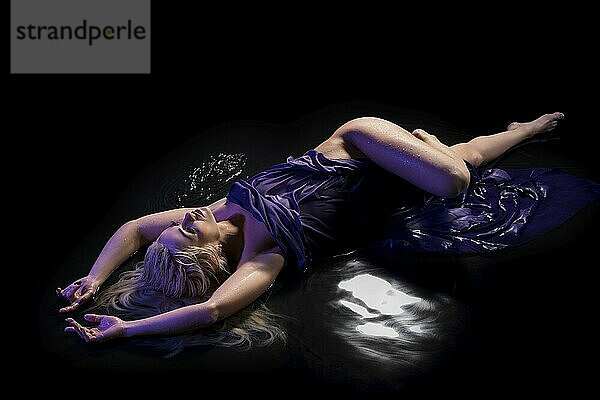 Sexy blonde wrapped in purple cloth lying in water in the dark room high angle view