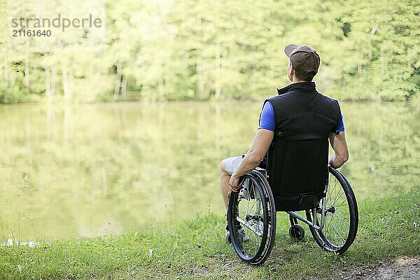 Happy and young disabled or handicapped man sitting on a wheelchair looking at beautiful lake in nature