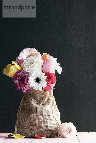 Canvas gift bag filled with multicolored roses  tulips and gerbera  on a pink table and a black background  surrounded by a rose and petals