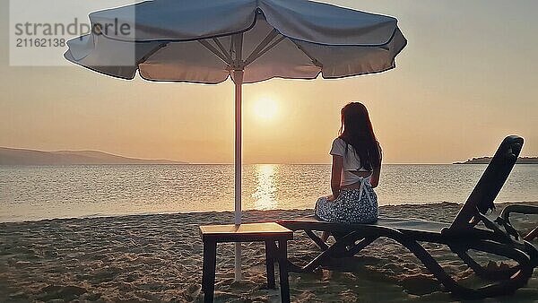 Ästhetische junge Frau auf einer Sonnenliege am Strand sitzen und beobachten die Morgendämmerung ein Sonnenbad mit Blick auf das Meer zu nehmen. Schöne Szene am Meer  Sommerurlaub Entspannung und Urlaub genießen Konzept