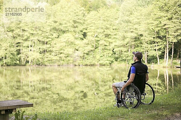 Happy and young disabled or handicapped man sitting on a wheelchair looking at beautiful lake in nature