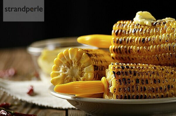 Wooden table with deep grilled sweet corn cobs under melting butter with plastic holder on dark background