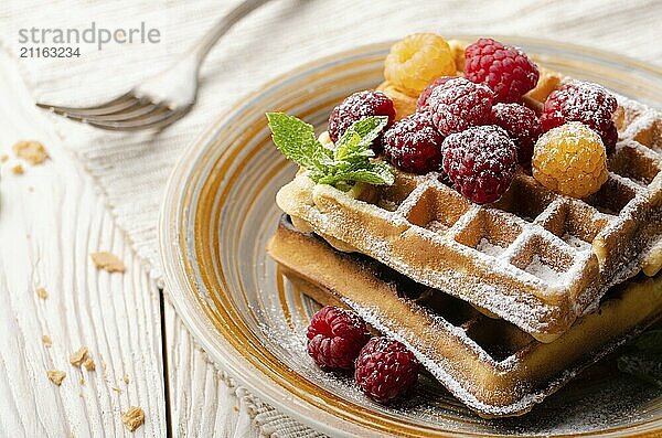 Belgian waffles served with raspberries and mint leaf dusted with powdered sugar on white wooden kitchen table