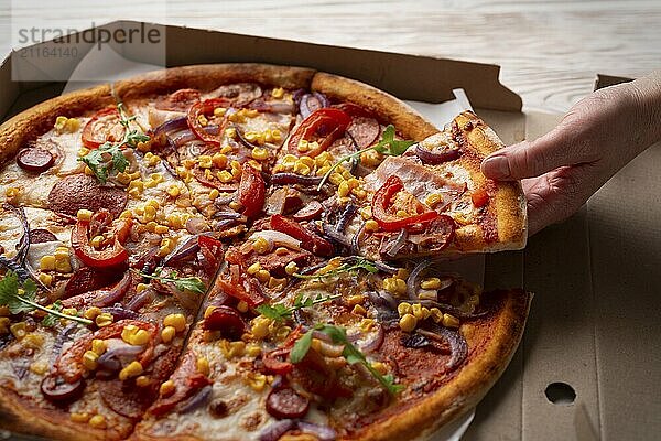 Human caucasian hand takes slice of large pizza from carton box on white wooden table