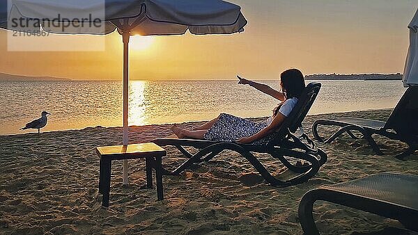 Junge Frau sitzt auf einer Sonnenliege am Strand und zeigt mit dem Zeigefinger auf den Sonnenaufgang am Horizont. Schöne Frau nimmt ein Sonnenbad am Meer. Sommerurlaub am Meer  Urlaub Genießen Konzept