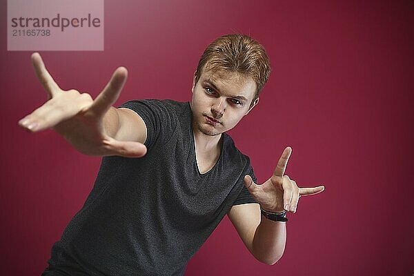 Young pathos Teenager boy wearing grey t-shirt over isolated pink background doing rock symbol with hands up. Music star. yo sign. cool sign