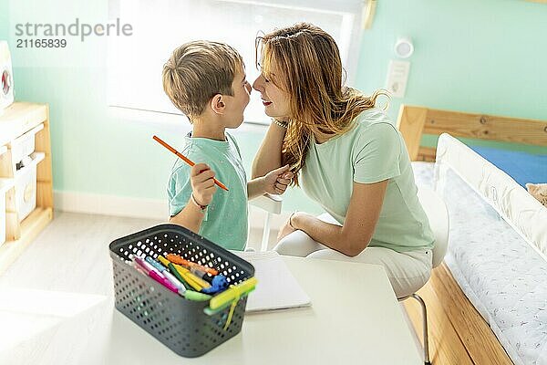 Tender mother and son rubbing noses while coloring sitting on the bedroom at home