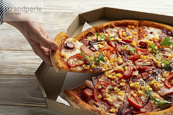 Human caucasian hand takes slice of large pizza from carton box on white wooden table