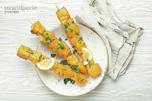 Gegrillte Lachsspieße gewürzt mit grüner Petersilie und Zitrone auf Keramikteller auf weißem Holztisch mit rustikalem Hintergrund von oben  gesunde Ernährung  Foodfotografie