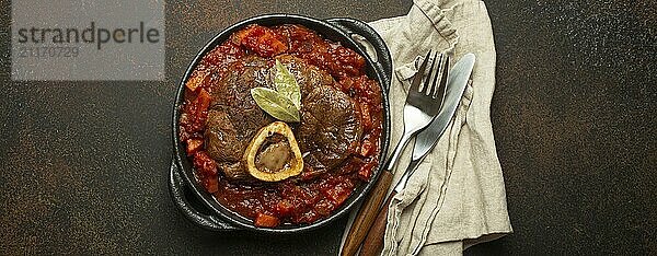 Traditionelles italienisches Gericht Ossobuco all Milanese aus geschnittener Kalbshaxe mit vegetarischer Tomatensoße  serviert in schwarzer Kasserolle von oben auf rustikalem braunem Hintergrund  Foodfotografie