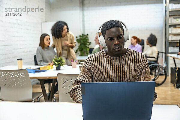 Afrikanischer Mann arbeitet mit Laptop und Kopfhörern in einem Coworking Center  während die Kollegen im Hintergrund reden