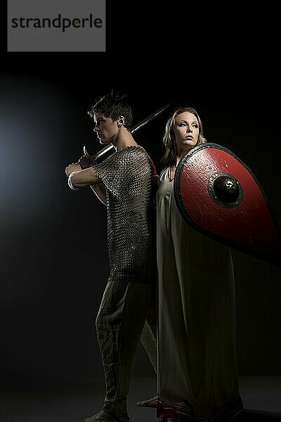 Young viking holding a sword and his beautiful wife with a shield staying back to back shot against black wall