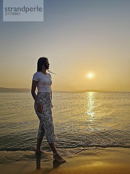 Ästhetische junge Frau zu Fuß barfuß im Wasser am Strand in der Morgendämmerung  beobachten die warmen Sonnenaufgang über dem Meer. Schöne Morgenszene am Meer  Sommerurlaub Entspannung Konzept