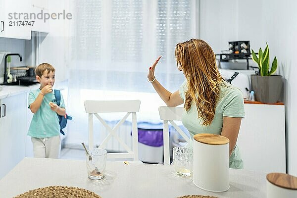 Mother waving goodbye to her son before he goes to school while sitting on the kitchen after having breakfast together