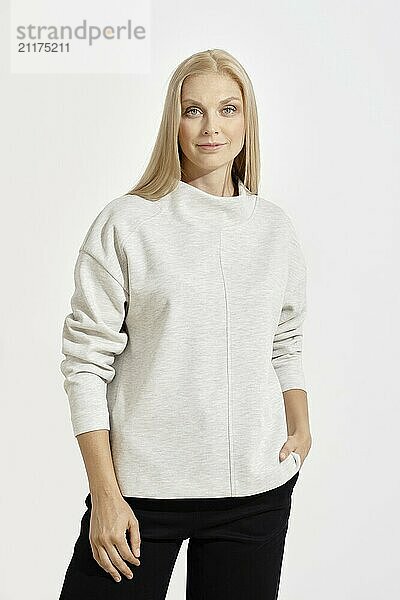 A woman with long blonde hair poses confidently in a light gray sweater  her hands resting casually in the pockets of her dark pants. The plain white backdrop emphasizes her relaxed demeanor