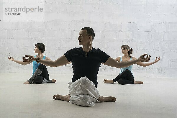 Male trainer and two women doing yoga exercises sitting on the floor view against brick wall