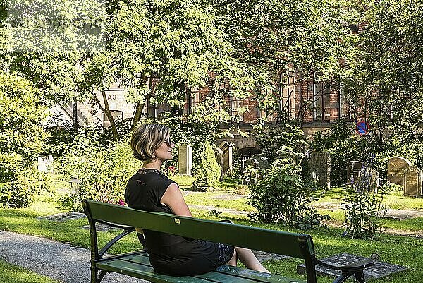 Dreißigjährige Frau in einem schwarzen Kleid auf einer Bank auf einem Friedhof im Sommer  Schweden  Europa