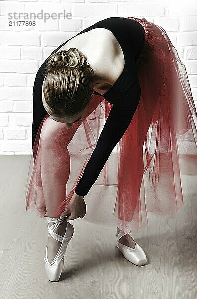 Young caucasian woman in ballet tutu lacing up her pointes on white brick wall background