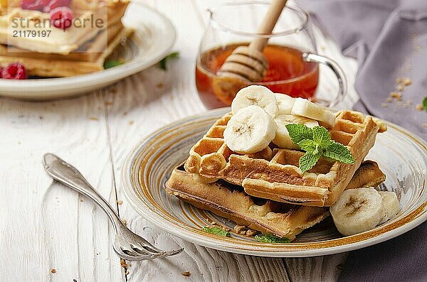 Belgian waffles served with banana and mint leaf on white wooden kitchen table with syrup aside