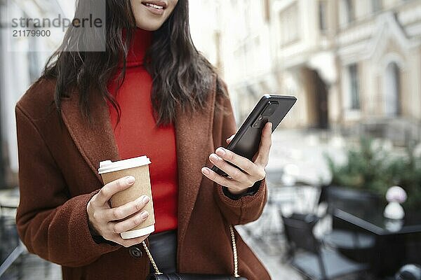 Smiling woman on the street using the phone and drinking coffee