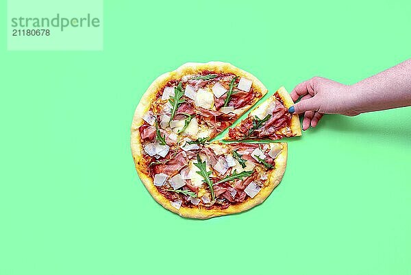 Top view with a homemade ham pizza isolated on a green background. Woman's hand taking a slice of pizza