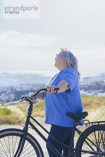 An overweight mature woman rides a black retro-style bicycle against a cloudy mountain backdrop with a village in the background