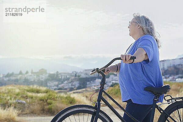 An overweight mature woman rides a black retro-style bicycle against a cloudy mountain backdrop with a village in the background