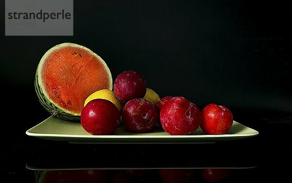 Water melon and plums and lemons oon a green plate with reflection on a dark background still life