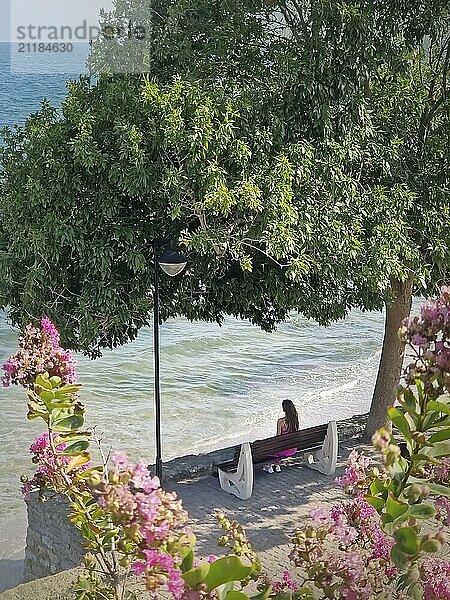 Junge Frau genießt einen schönen Sommertag auf einer Bank unter einem grünen Baum mit Blick auf das Meer sitzen und genießen die Brise. Seaside genießen Konzept  friedliche und entspannte Atmosphäre