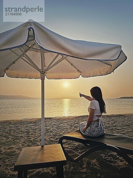 Junge Frau sitzt auf einer Sonnenliege im Ferienort Sunny Beach in Bulgarien und zeigt mit dem Zeigefinger auf den Sonnenaufgang über dem Horizont. Schöne Frau nimmt ein Sonnenbad am Meer in der Nähe von Nessebar. Sommerurlaub am Meer