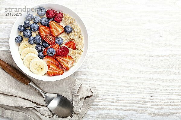 Oatmeal porridge with fruit and berries in bowl with spoon on white wooden background table top view  homemade healthy breakfast cereal with strawberry  banana  blueberry  raspberry. Space for text  food photography