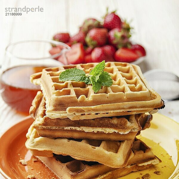 Pile of belgian waffles on white wooden kitchen table with strawberries and syrup aside