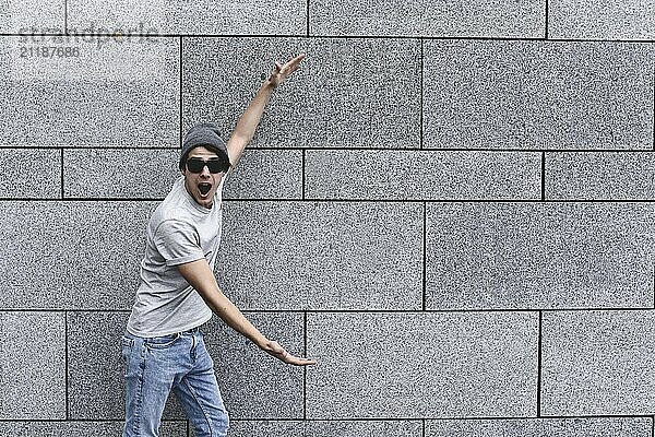 Cheerful guy dressed in gray t-shirt  sunglasses and hat at the street  show a big sale  or nice offer. Advertise concept. People  travel and tourism  man with Sunglasses on city street and guy show with his hands something over gray wall background