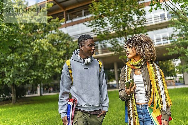 Frontal view of two multiethnic students chatting and using mobile walking out the university