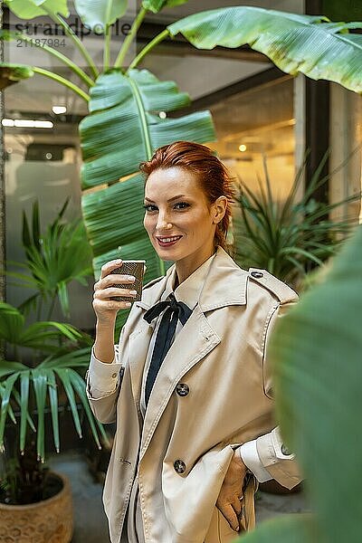 Vertikales Porträt einer eleganten Frau  die am Eingang des Büros steht und Kaffee trinkt