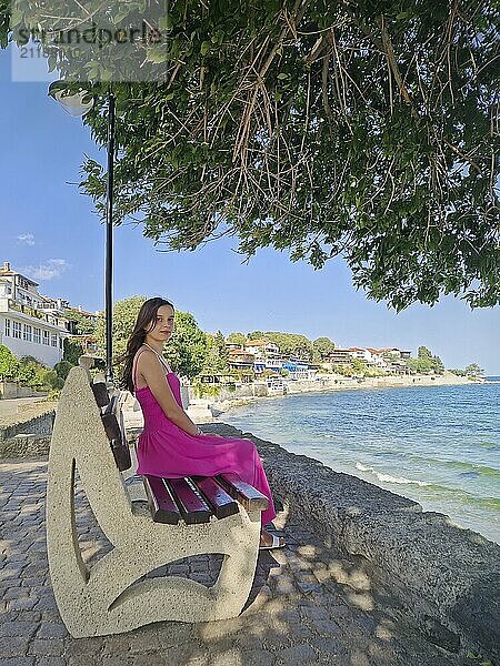 Schöne junge Frau in rosa Kleid  sitzt auf einer Bank und beobachtet das Meer und die Altstadt von Nessebar Küste. Sommerurlaub Reise Szene in Bulgarien