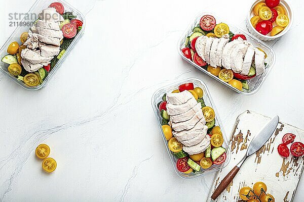 Healthy meal prep for lunch  diet boiled chicken fillet with vegetables salad in lunch boxes containers top view on white marble kitchen table background with knife and cutting board. Space for text  food photography