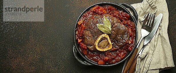 Traditionelles italienisches Gericht Ossobuco all Milanese aus geschnittener Kalbshaxe mit vegetarischer Tomatensoße  serviert in schwarzer Kasserolle von oben auf rustikalem braunem Hintergrund  copy space  Food Photography