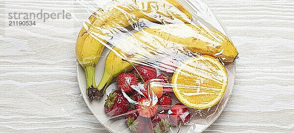 Assorted verschiedene Früchte und Beeren auf weißem Teller in Kunststoff eingewickelt klar transparente Folie von oben auf weißem Holz rustikalen Tisch Hintergrund  Lebensmittel Fotografie