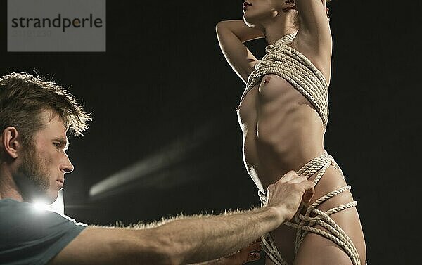 Man tying nude woman with rope cropped low angle view in dark room