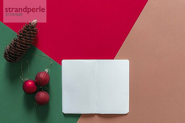 Christmas concept in a minimal style with a white empty notebook  red Christmas balls  and a pine cone  on a red  green and orange background