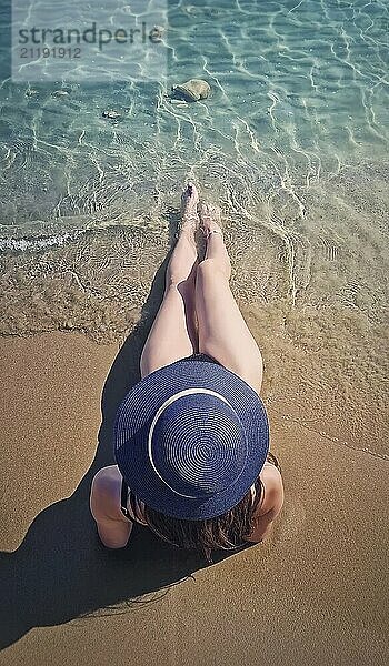 Rückansicht einer jungen Frau mit blauem Sonnenhut  die am Strand sitzt und die Beine ins Meer streckt. Das klare Wasser umspült sanft ihre Füße. Gelassene und entspannende Sommerurlaubsszene