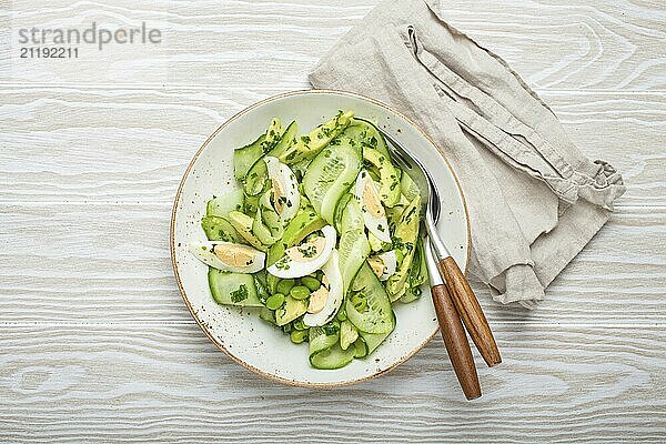 Gesunde grüne Avocado Salatschale mit gekochten Eiern  Gurkenscheiben  Edamame Bohnen  Olivenöl und Kräutern auf Keramikteller Draufsicht auf weißem Holztischhintergrund  Foodfotografie