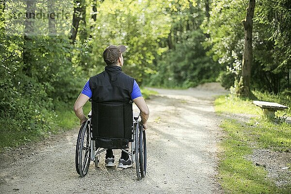 Happy and young disabled or handicapped man sitting on a wheelchair in nature turning wheels on a walking road at a beautiful sunny day