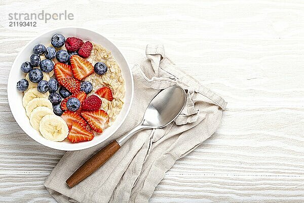 Haferflockenbrei mit Obst und Beeren in der Schüssel mit Löffel auf weißem Holzhintergrund Tischansicht  hausgemachte gesundes Frühstück Müsli mit Erdbeere  Banane  Heidelbeere  Himbeere. Platz für Text  Food Fotografie