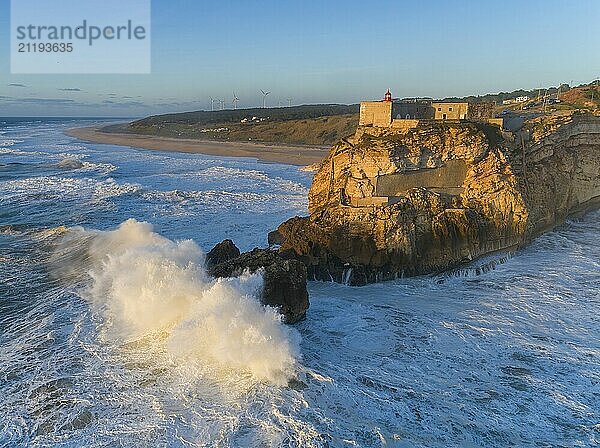 Aerial view of lighthouse on a cliff with a fortress on the coast of the Atlantic ocean with big waves at sunset in Nazare  Portugal  Europe