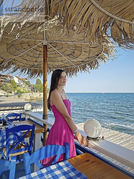 Gelassene Szene mit einer hübschen jungen Frau in einem rosa Kleid  die unter einem alten Sonnenschirm auf einer Cafe Lodge mit Blick auf die Küste steht und das blaue Meerwasser bewundert. Entspannung und Frieden Reisekonzept