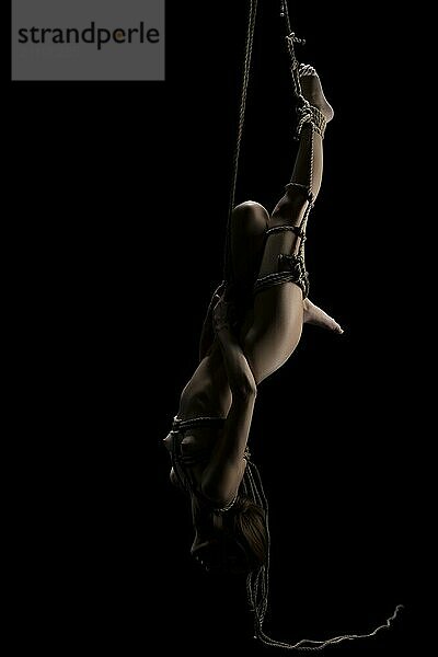 Naked beautiful blonde tied up with a rope hanging upside down isolated shot on black background