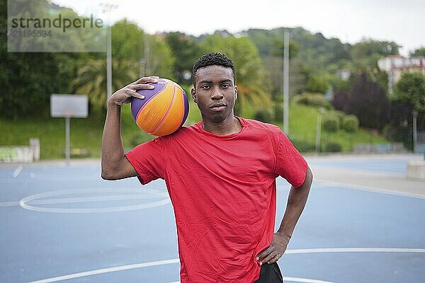 African man posing with basketball ball on shoulder in an urban basketball outdoors court