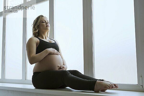 Young pregnant blonde in sportswear sitting on a window sill shot